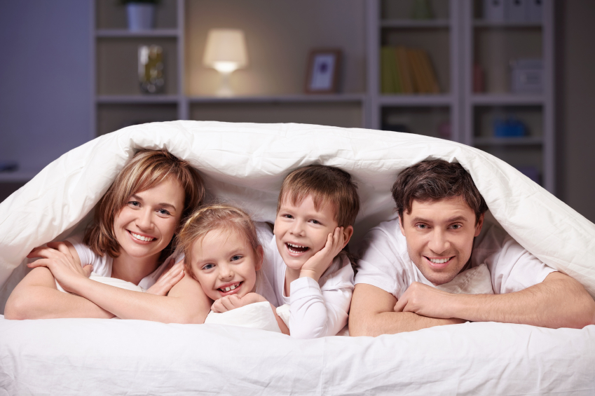 Young family smiling and posing under blankets with lamp lit in background.