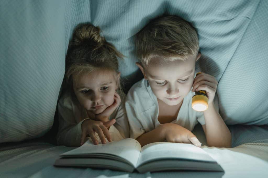 Two young children hiding under blanket and using flashlight to read a book.