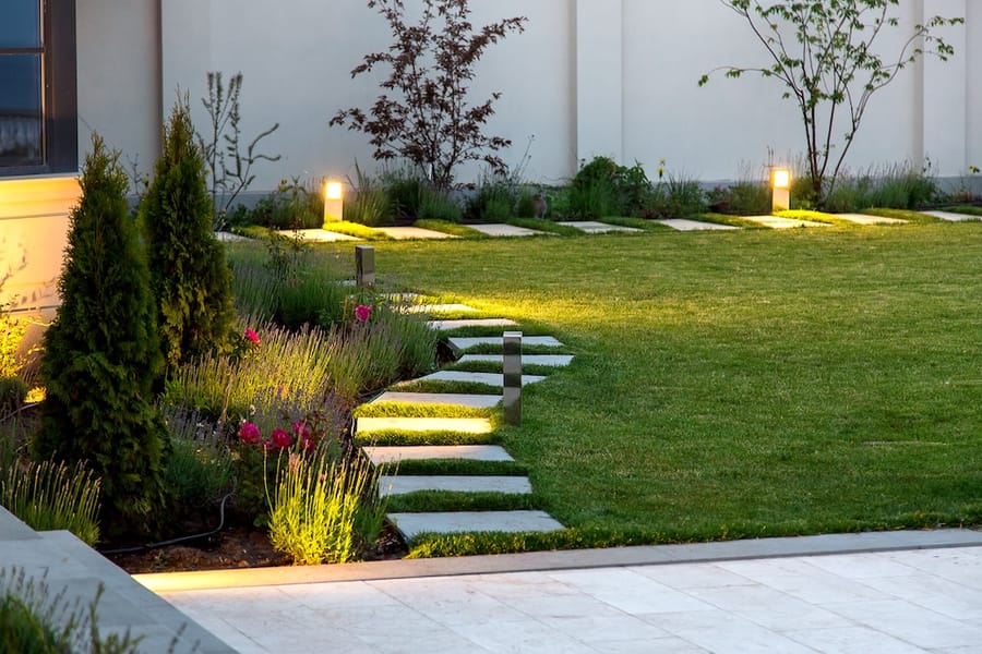 A stone pathway and garden beds illuminated by warm LED bollard lights, showcasing a professional landscape lighting installation by Preferred Home Services.