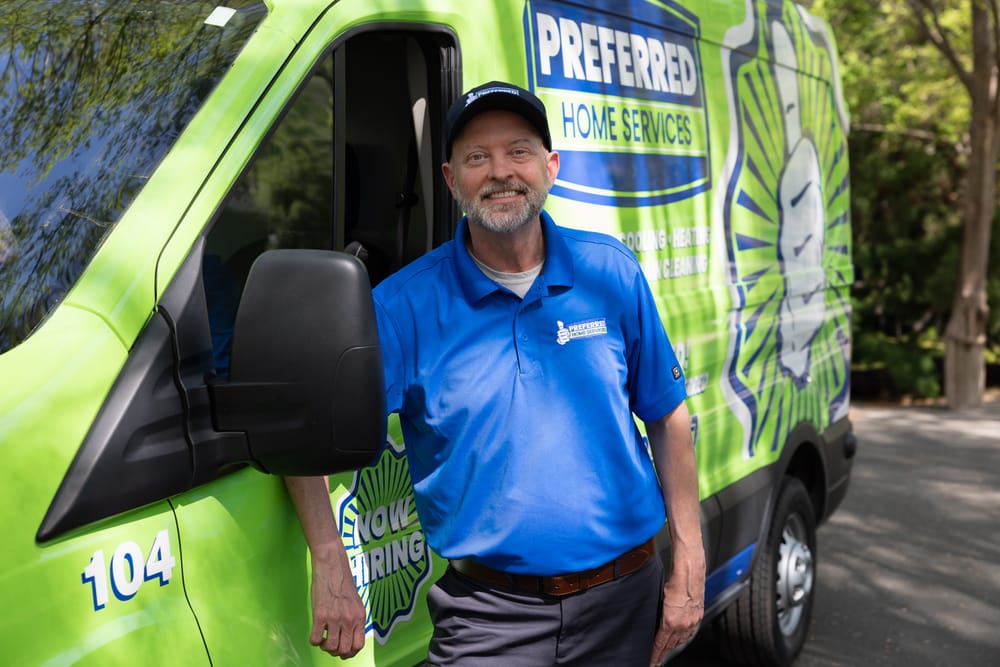 A friendly Preferred Home Services plumber waves from the driver's seat of a green service van, arriving at a customer's home to provide professional shower and bath repair services.
