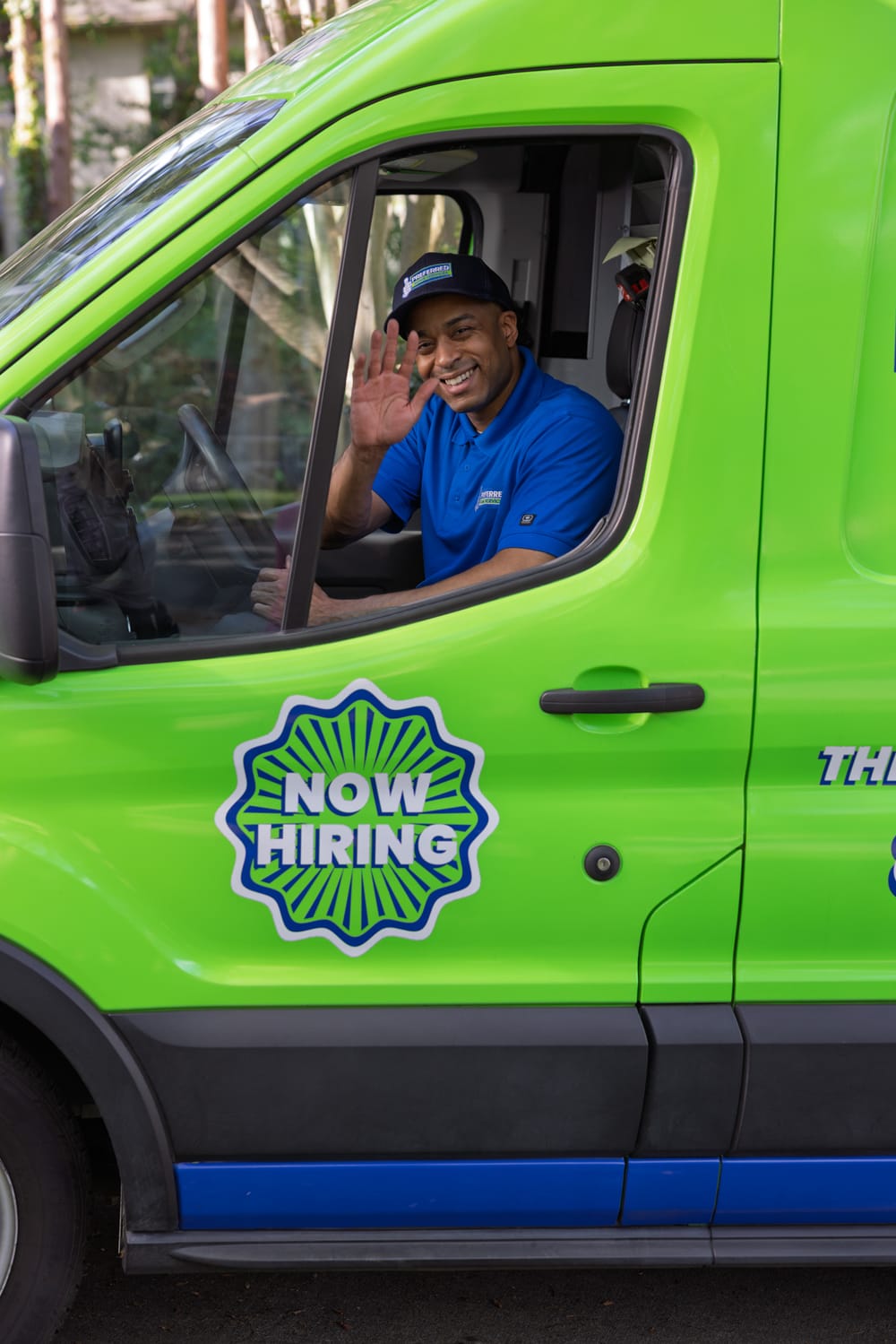 A friendly Preferred Home Services technician waves from a green service van while arriving at a residence to perform professional water heater repair or installation.