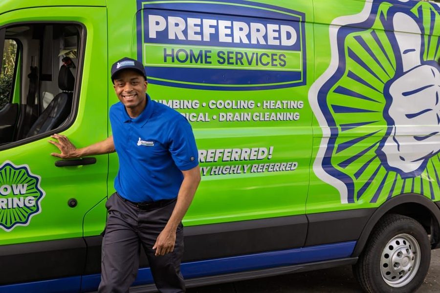 Smiling service technician in a blue polo stands beside a bright green Preferred Home Services van with its door open.