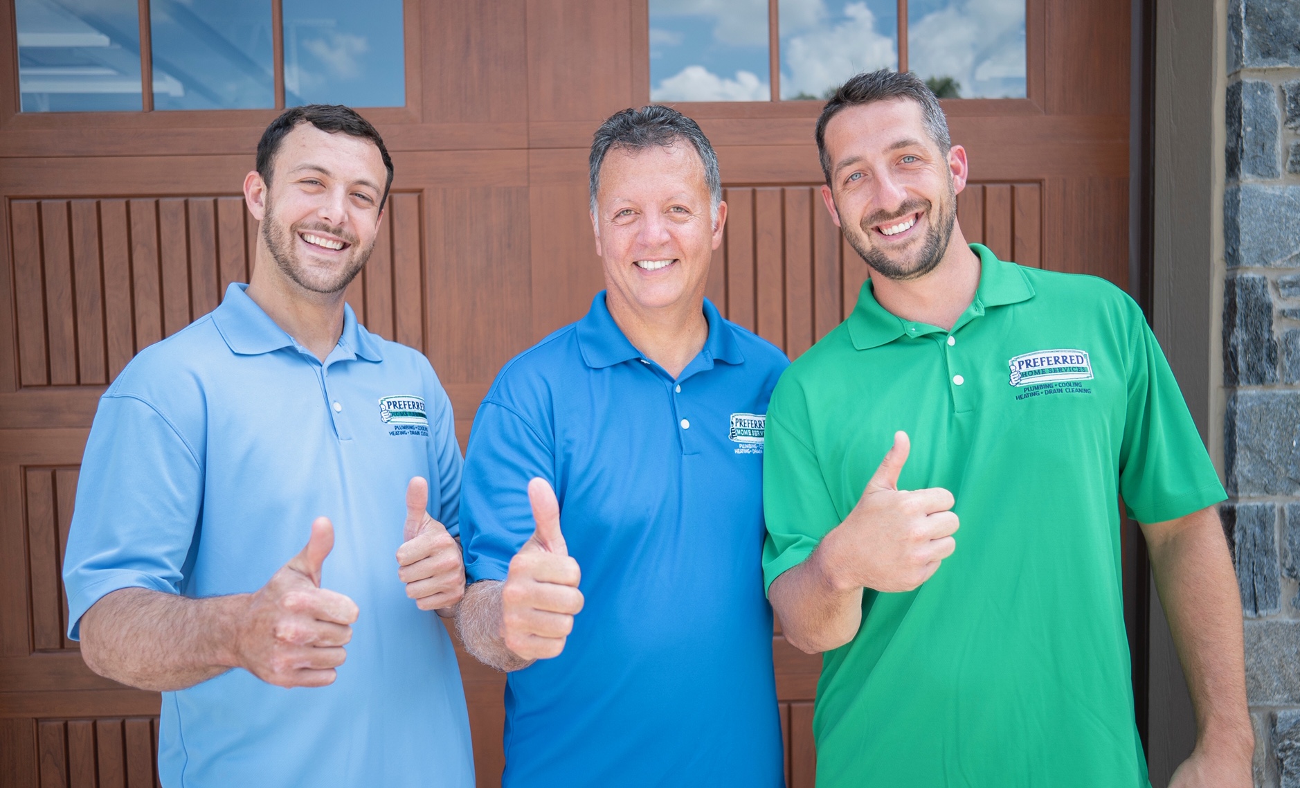 Three smiling men in blue and green polo shirts give thumbs up in front of a wooden door building façade.