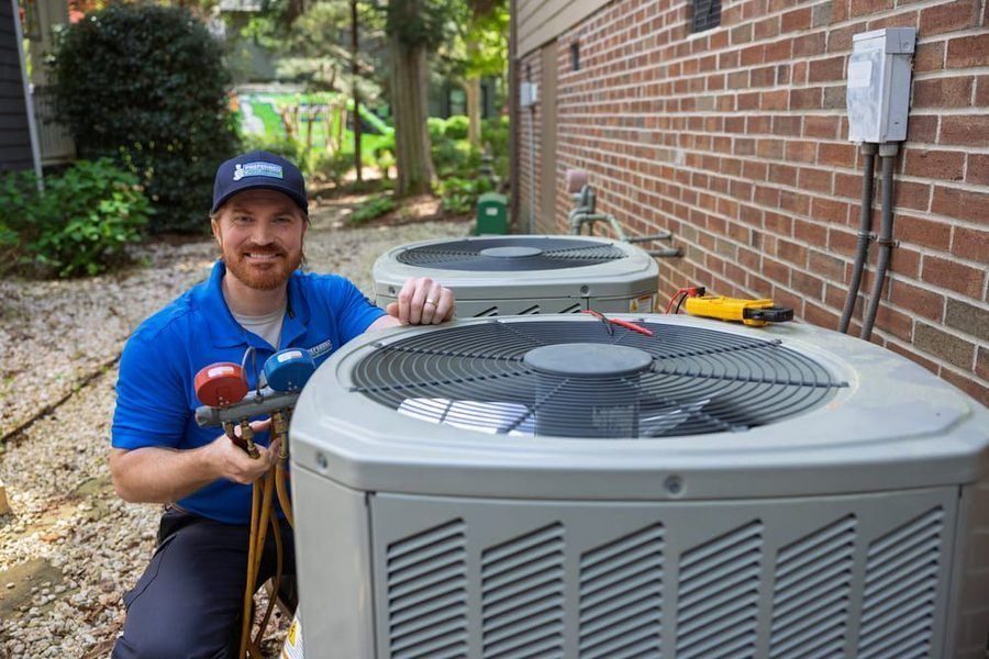 HVAC technician in a blue polo and cap inspecting two outdoor air conditioner units with refrigerant gauges outside a brick building