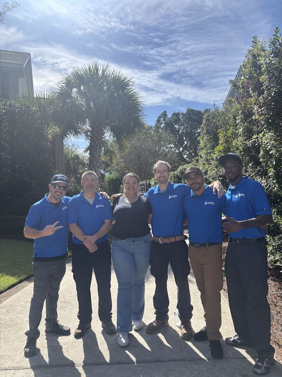 Six people posing together on a sunny residential sidewalk; five men in blue shirts and a woman in a black top, palm trees in the background.
