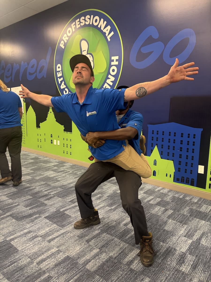 Man in a blue polo stretches arms wide while another person supports him from behind, indoors with a cityscape mural on the wall.
