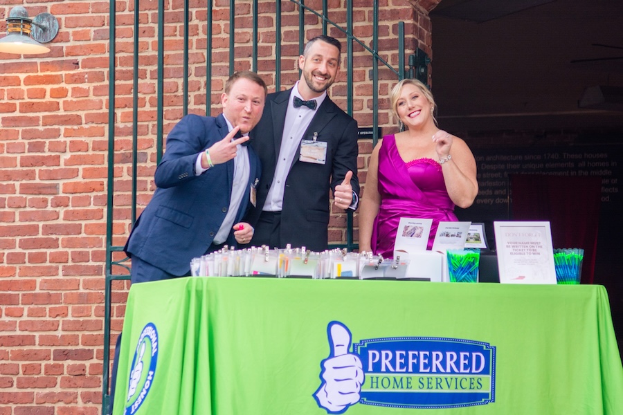 Three people pose cheerfully at a green Preferred Home Services booth with brochures and cups in front of a brick wall background