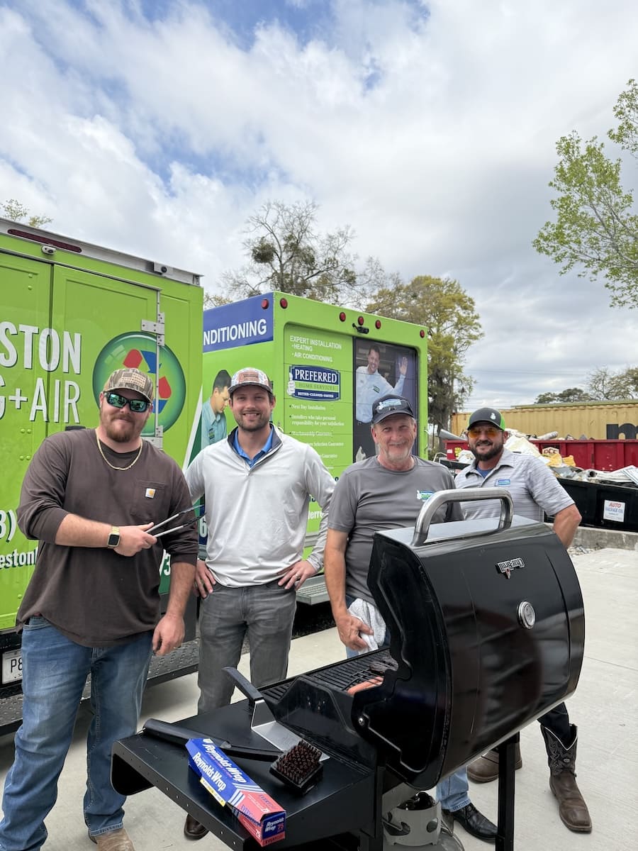 Four men stand beside a large black grill in a parking lot, with green service trucks in the background.
