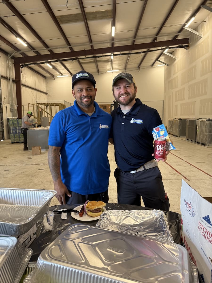 Two coworkers pose and smile in a warehouse; one holds a can of Dr Pepper and chips, with a burger on a plate on a foil-covered table in front of them.