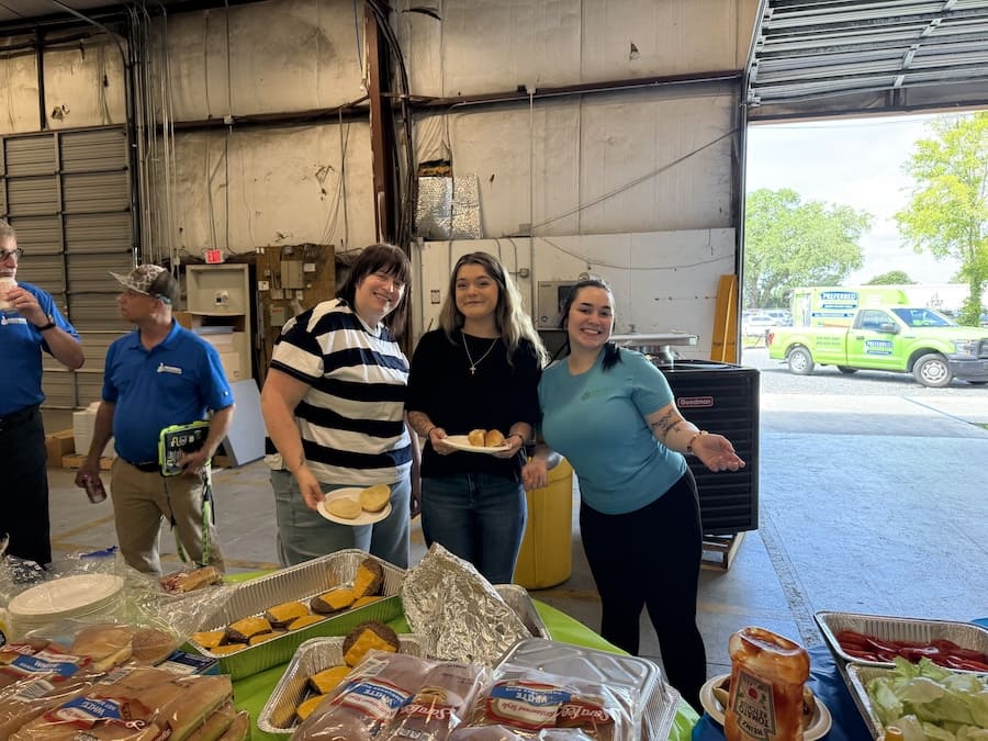 Four people stand inside a large open garage, smiling and holding plates of buns and small sandwiches around a table of food. The group appears to be at a casual community event or gathering.