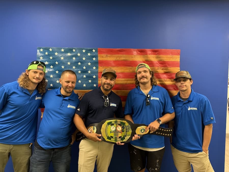 Five men in blue polo shirts pose together, holding a green-and-yellow championship belt in front of a United States flag.