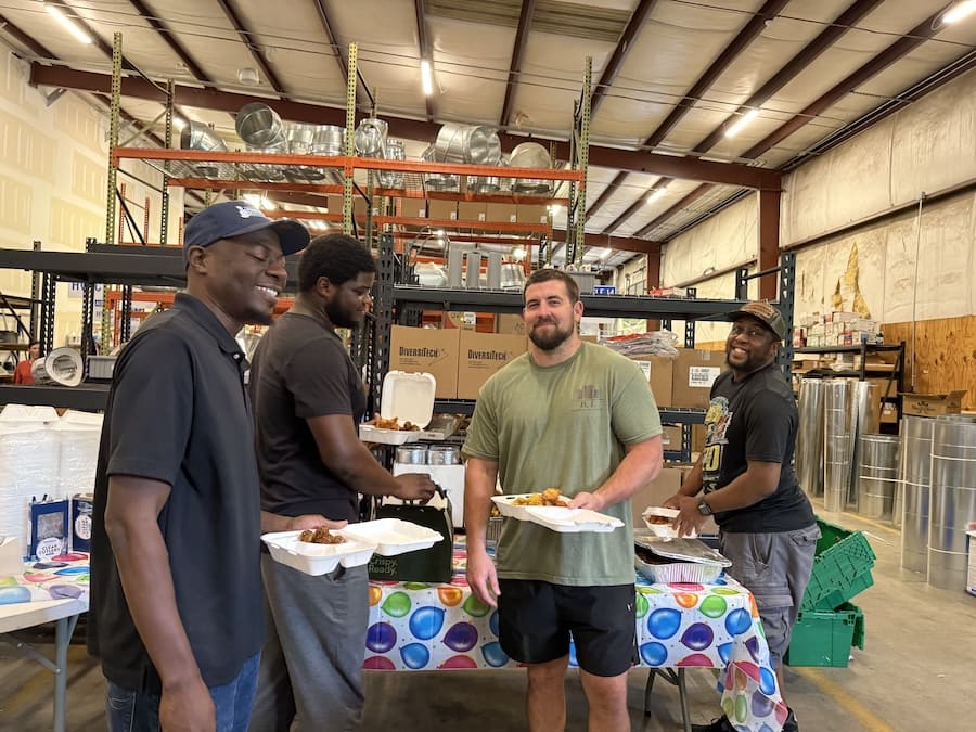 Four men stand in a warehouse holding takeout plates at a table covered with a colorful tablecloth, smiling and chatting while eating.