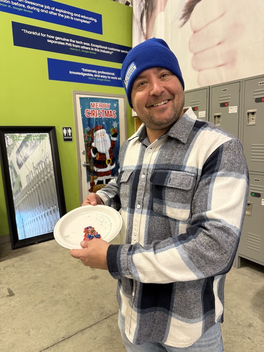 Smiling man in a blue beanie holds a white plate with a colorful craft, standing near lockers with a Christmas-decorated door in the background.