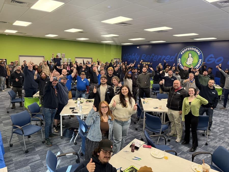 Large group of coworkers in a bright meeting room, many giving thumbs up around tables with snacks and papers nearby.