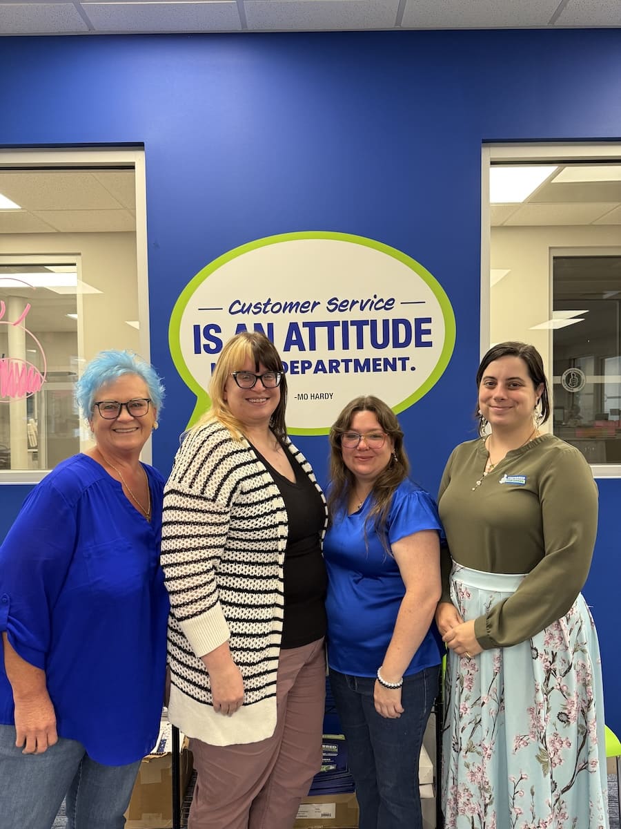 Four women pose indoors in front of a blue wall with a sign that reads 'Customer Service is an Attitude'—a friendly team photo.