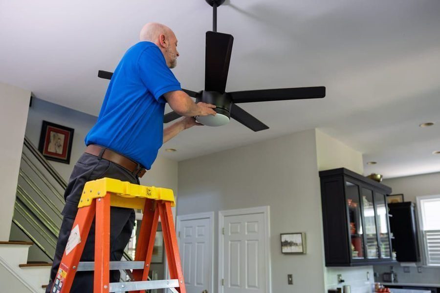 Workman on a yellow-top ladder installing a ceiling fan with black blades in a bright kitchen.