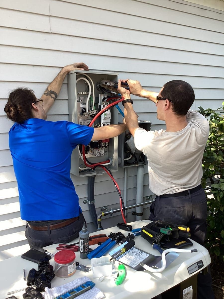 Two workers repair an outdoor electrical panel on the side of a house with tools laid out on a table nearby.