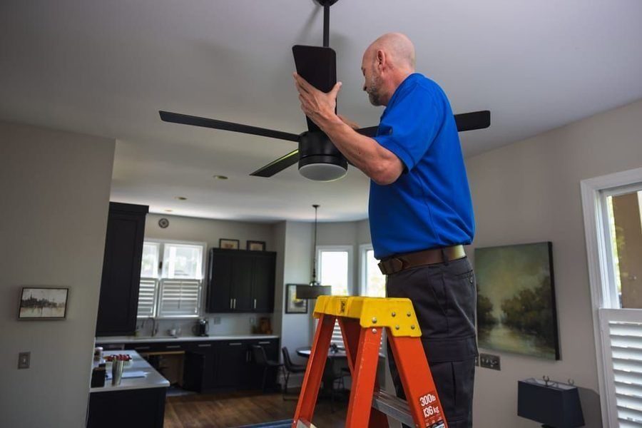 Worker on an orange ladder installing a ceiling fan in a modern kitchen/living area.