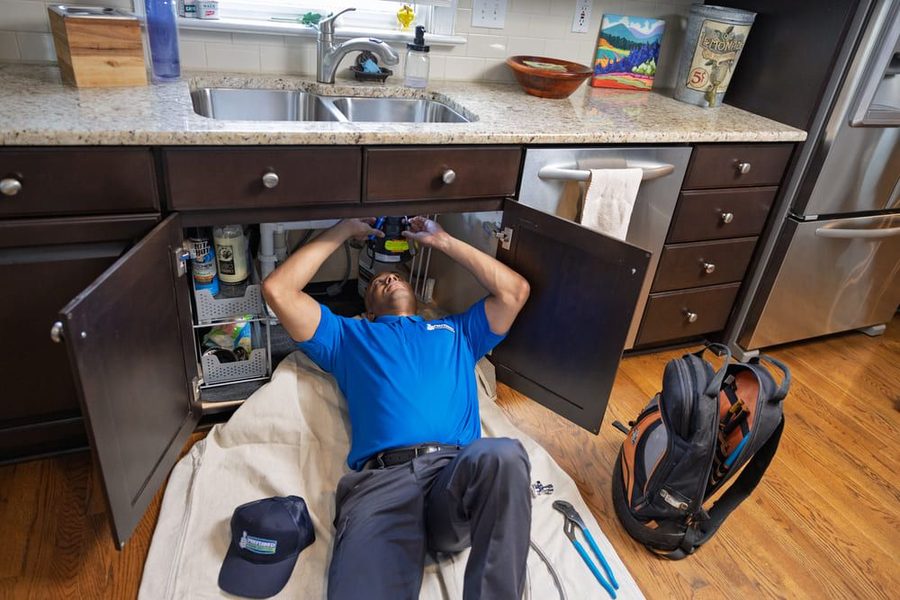 Plumber in a blue shirt works under a kitchen sink with cabinet doors open and tools nearby.