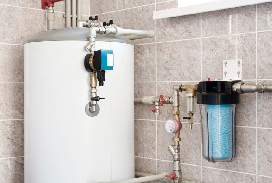 Residential water heater with blue control valve and attached piping in a tiled utility room, showing filtration components to the right and a pressure gauge on the line.