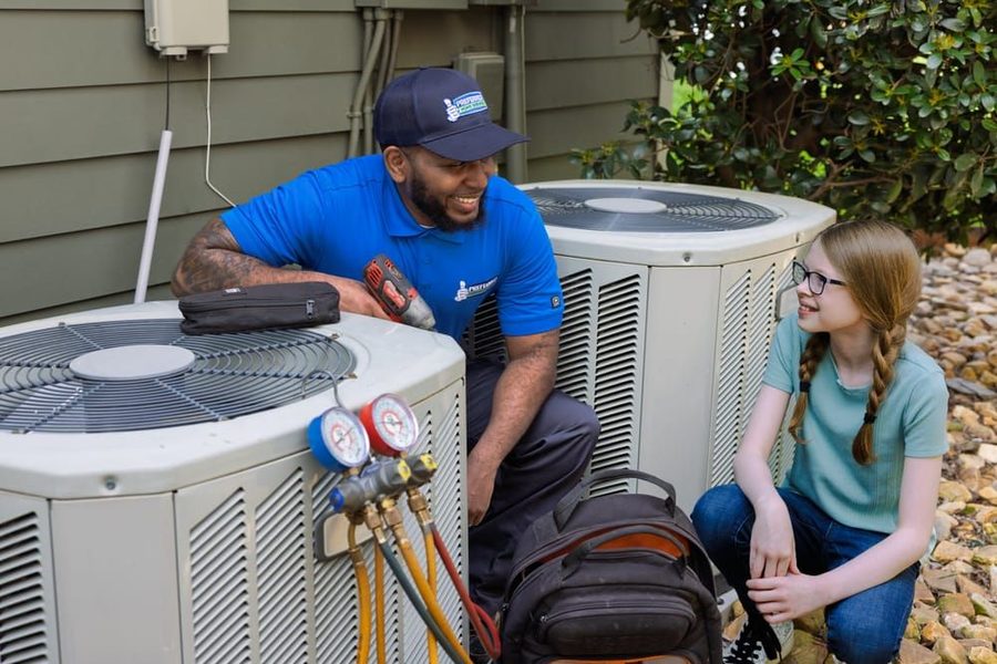 HVAC technician in a blue polo explains gauge readings to a smiling girl beside outdoor air conditioner units.