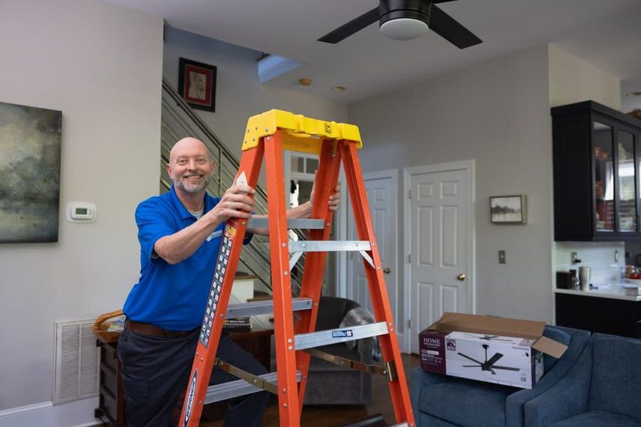 Man in a blue polo stands beside an orange ladder in a living room, preparing to install a ceiling fan above him.