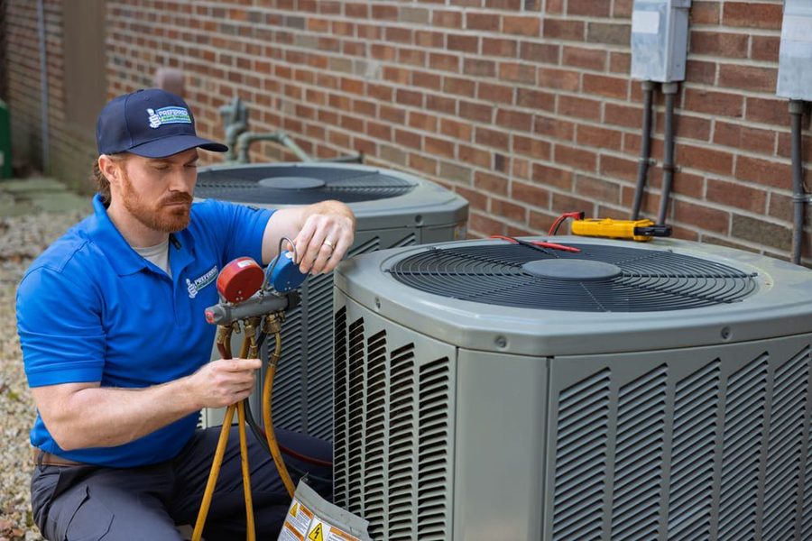 HVAC technician in a blue polo checks refrigerant pressure on an outdoor air conditioner unit with a manifold gauge set.