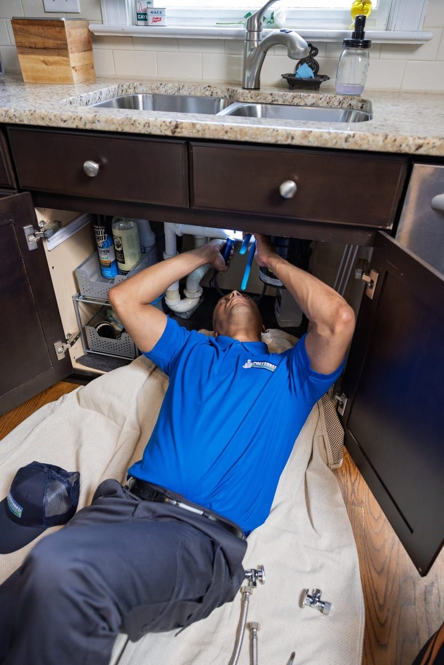 Plumber lying under a kitchen sink, working on exposed pipes with tools nearby under cabinet.