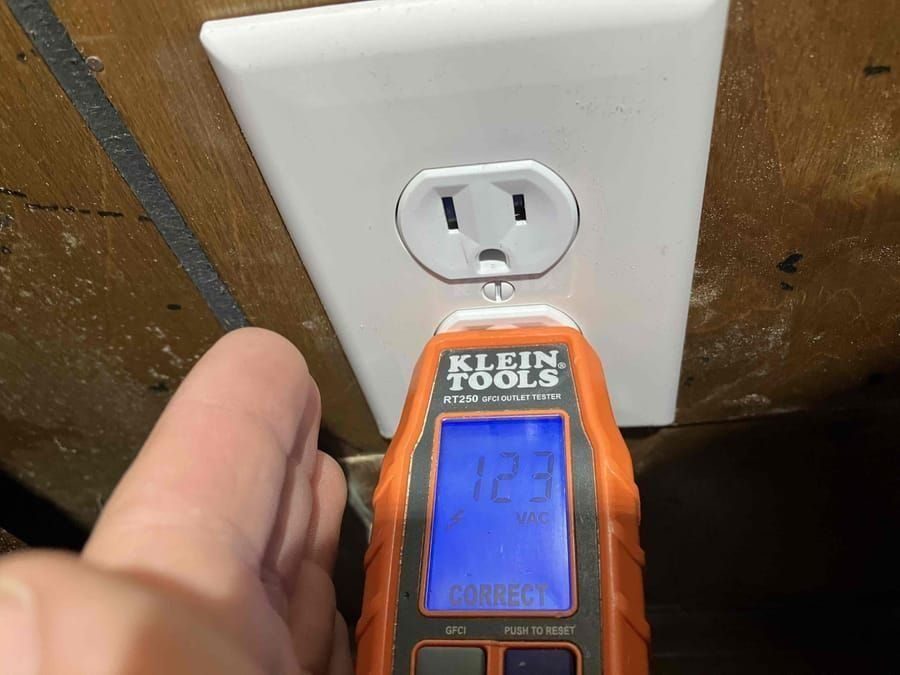Hand holding an orange Klein Tools outlet tester plugged into a wall outlet, displaying 123 VAC and 'CORRECT' on the screen.