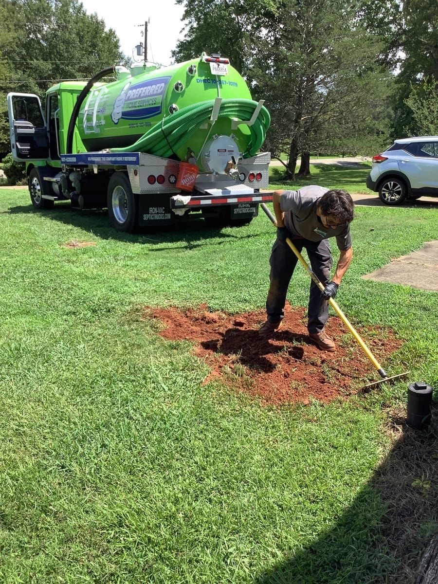 Technician with gloves digging a hole in a lawn beside a bright green septic service truck with hoses in the background.