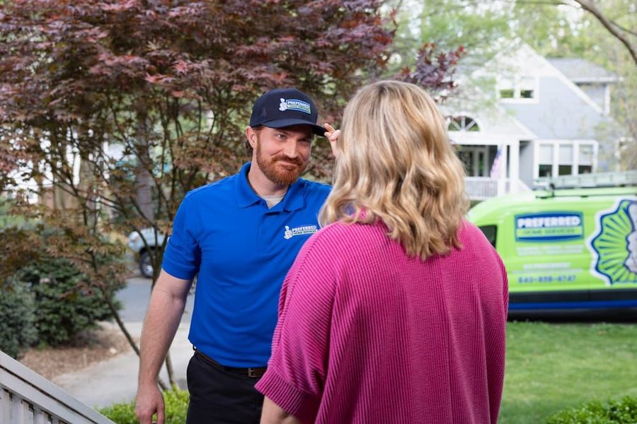 A man in a blue polo shirt speaks with a blonde woman on a doorstep, with a lime-green service van in the yard in the background.