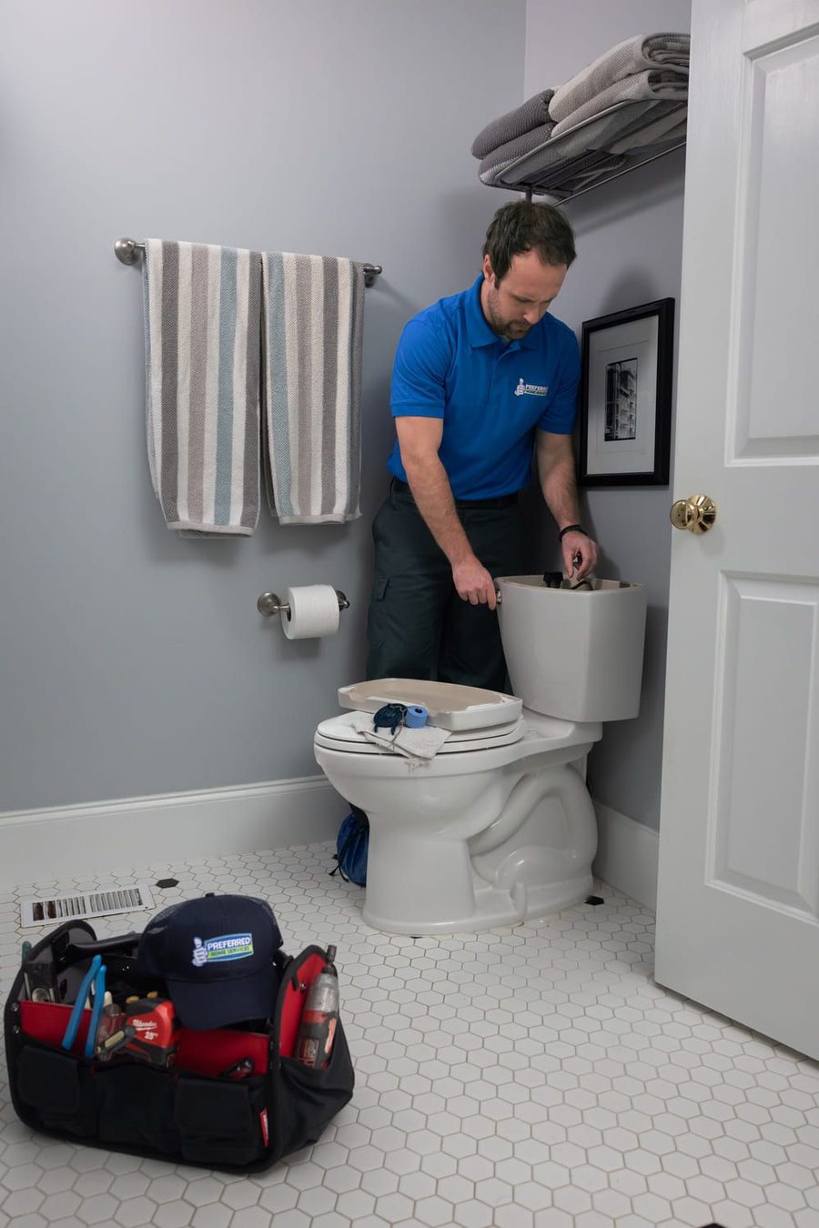 Plumber in a blue polo shirt works on a toilet tank in a bathroom; a tool bag sits on the tiled floor nearby.