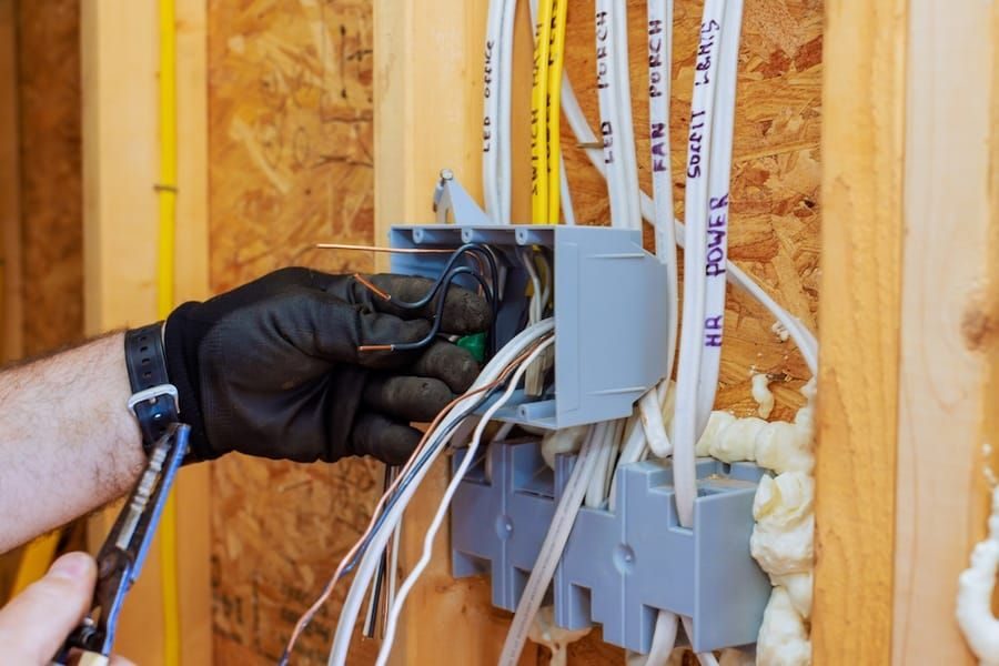 Gloved hand wiring a gray electrical junction box with multiple cables in a wooden wall frame.